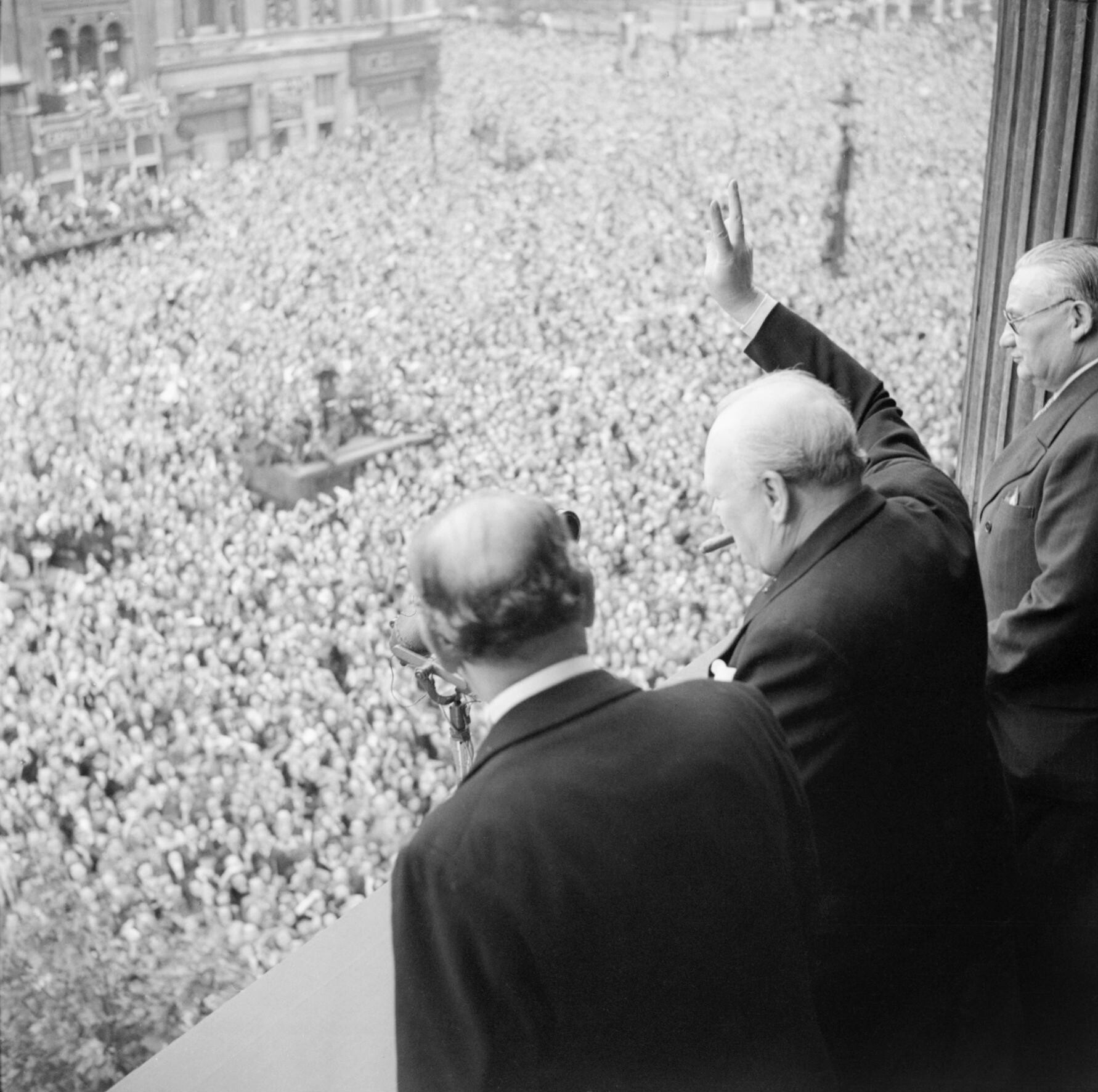 Winston Churchill mává davům ve Whitehallu v Londýně během oslav Dne vítězství (VE Day), 8. května 1945. Z balkonu ministerstva zdravotnictví ukazuje své slavné gesto „V jako vítězství“ v den, kdy v rozhlasovém vysílání oznámil národu, že válka s Německem byla vyhrána. Po Churchillově levici stojí sir John Anderson, kancléř pokladu (ministr financí). Po Churchillově pravici je Ernest Bevin, ministr práce.
